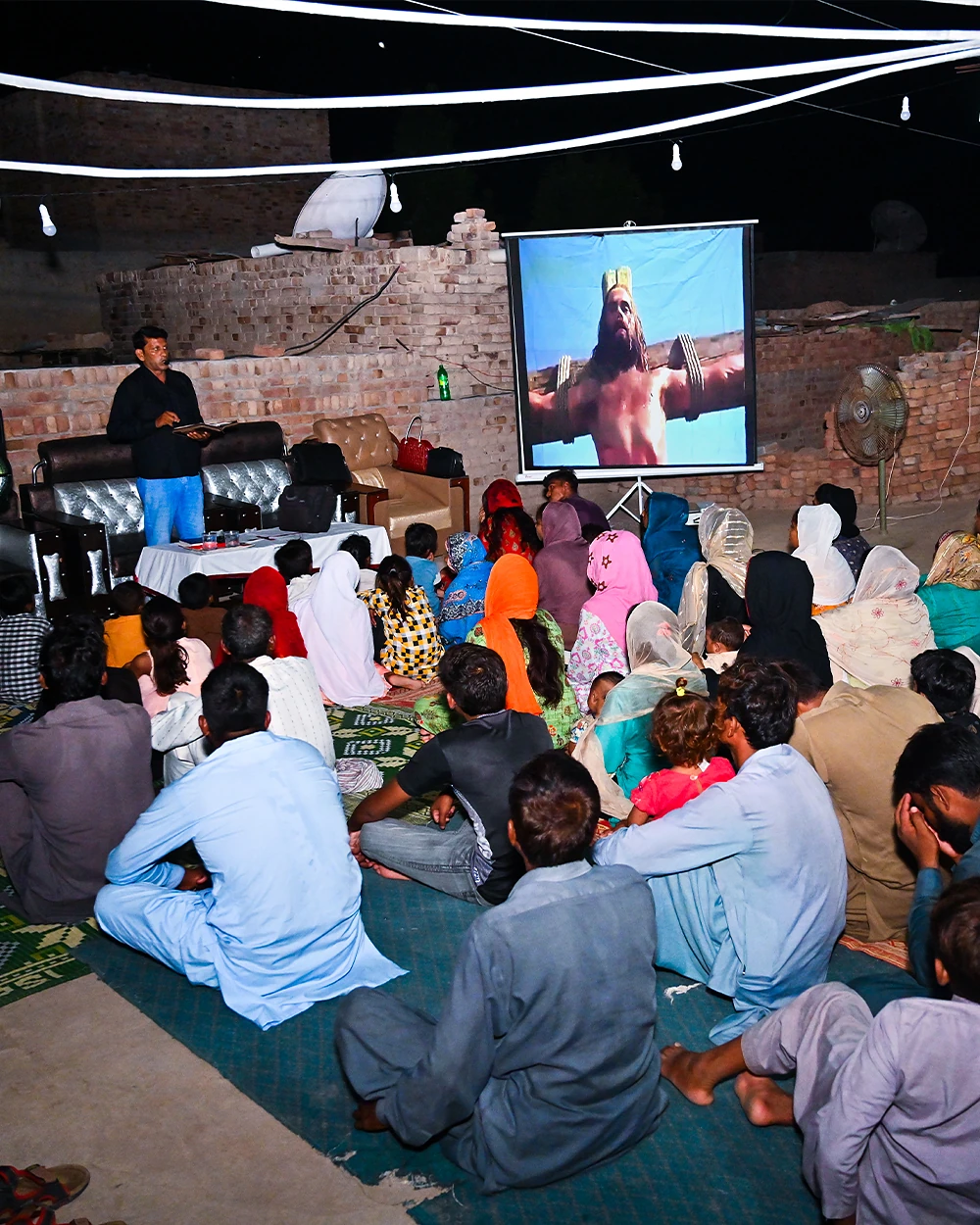 A village seated on the ground watching the Jesus Film projected in a courtyard after dark